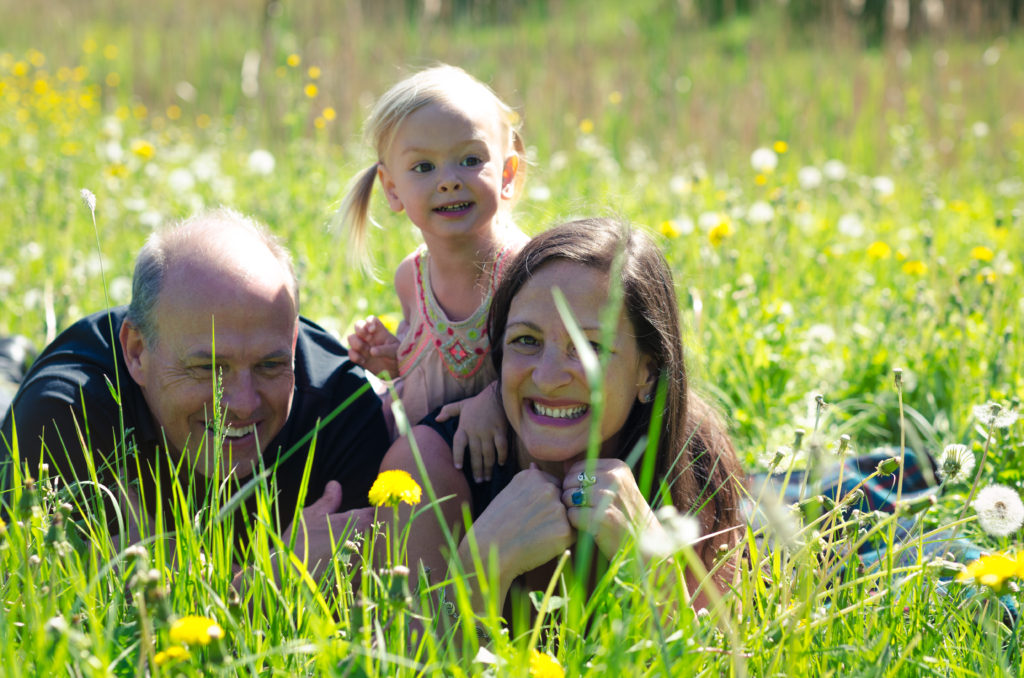mperfect Parenting Podcast: Mats, Ella and Ariel Andersson in Wishing Meadow, Prague on a sunny day. Parents laying stomach down side by side and 2 year old daughter sitting on mom's back; all smiling in the grass between flowers.